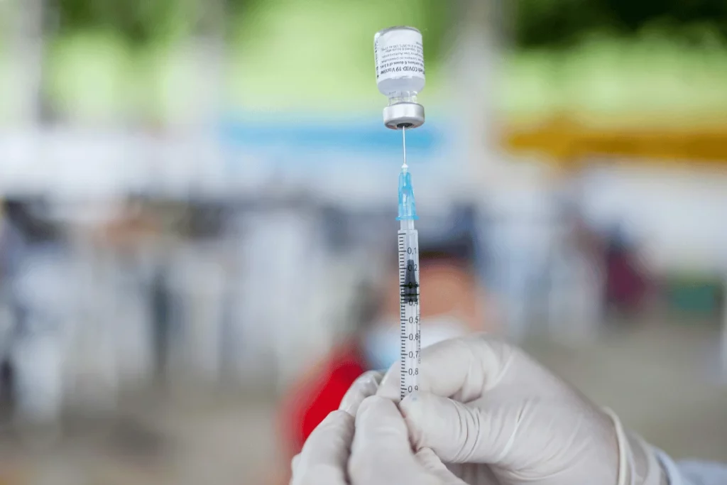 A gloved hand holds a syringe filled with vaccine fluid, poised to administer an injection in a blurred vaccination setting.