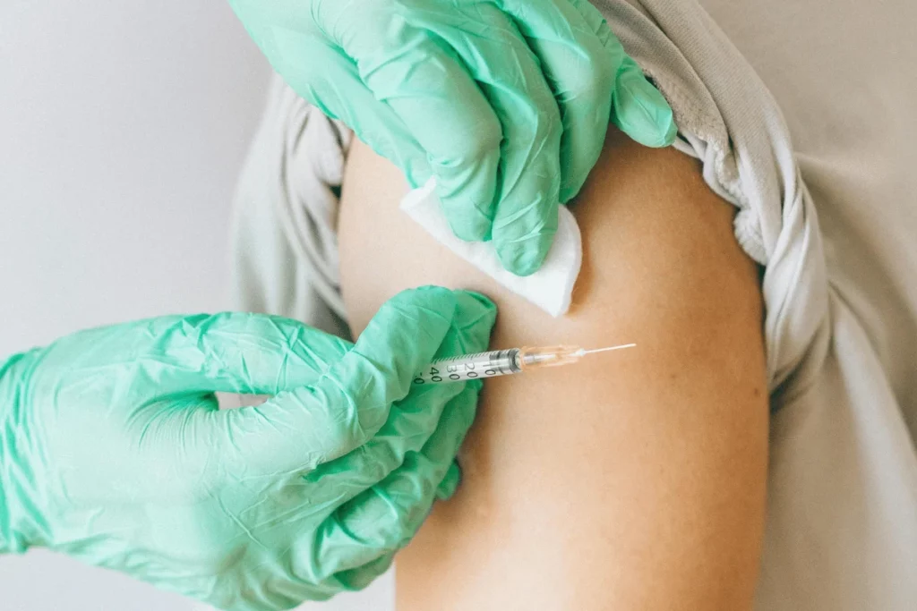 A healthcare worker in green gloves administers a vaccine with a syringe to a patient's arm, preparing with a cotton swab.