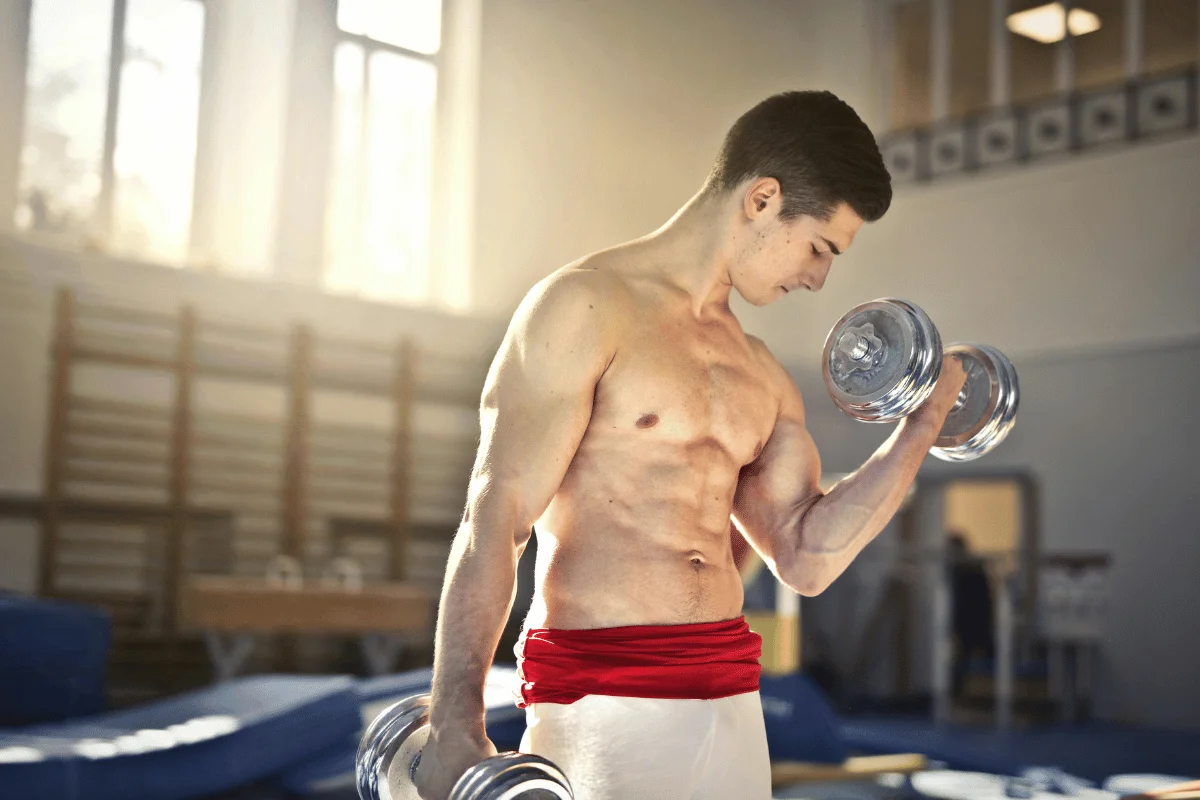 A muscular man in athletic attire lifts dumbbells in a sunlit gym, showcasing strength and determination.