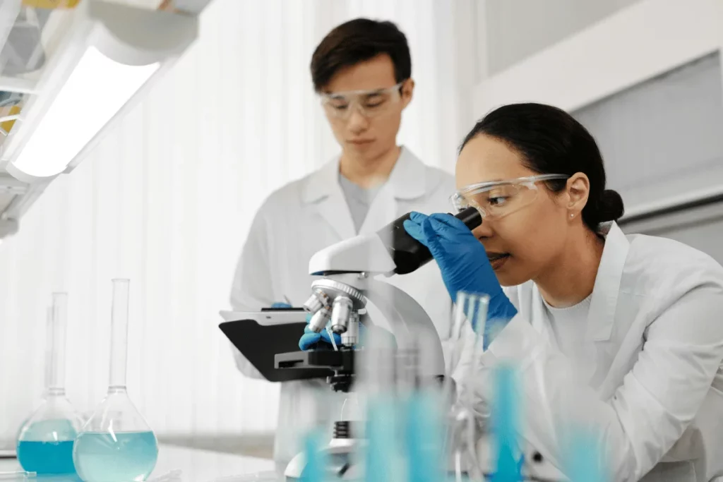 Female scientist using a microscope with a male colleague reviewing data in a laboratory.