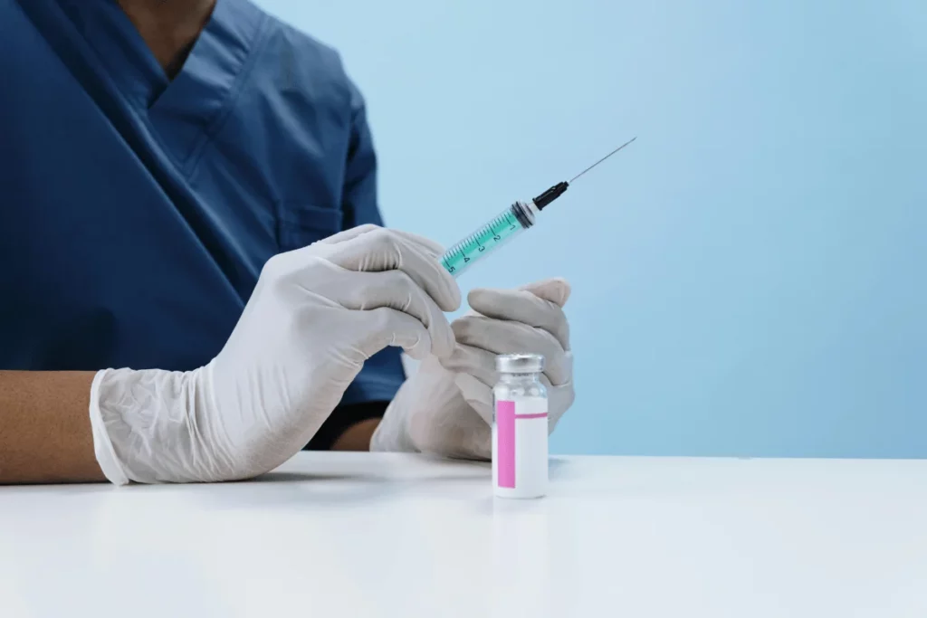 A healthcare professional in gloves holds a syringe ready to draw from a vial on a white surface against a light blue background.