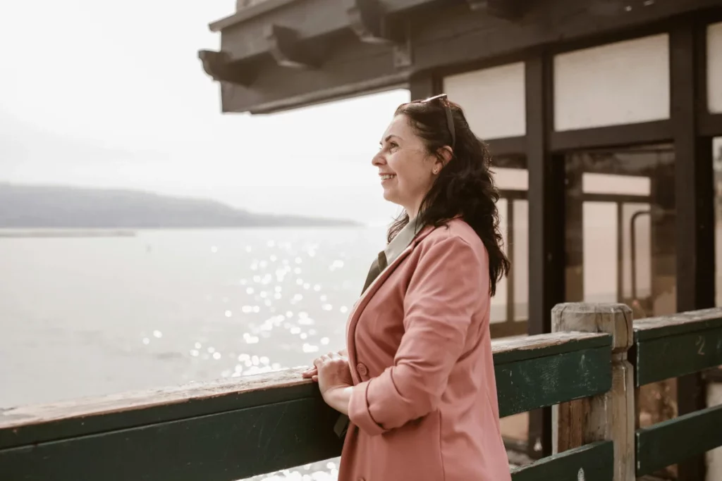 Smiling woman in a pink jacket enjoys the view while standing on a pier, looking at the peaceful water.