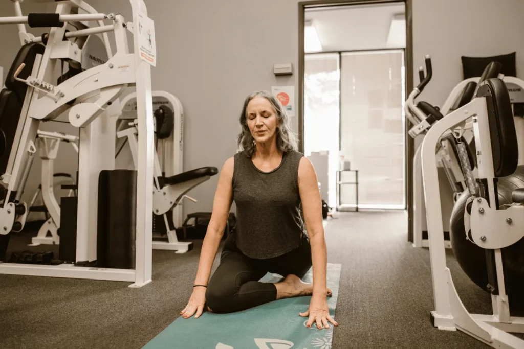 Mature woman practicing yoga in a gym setting, performing a stretching pose on a yoga mat.