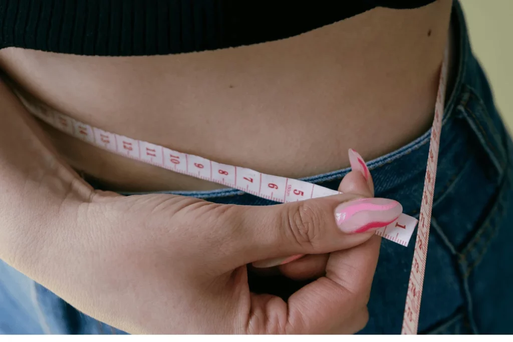 A woman measures her waist with a tape measure while wearing jeans and a black crop top, tracking her fitness progress.