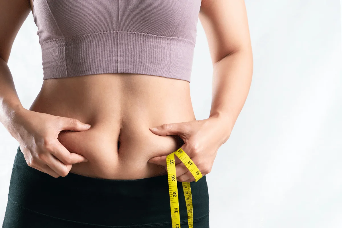 A woman in a purple crop top is measuring her waist with a yellow tape measure, looking focused on her fitness journey.