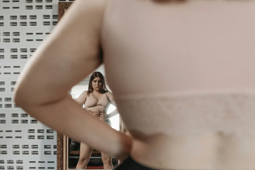 A woman in a beige bra looking at her reflection, highlighting body positivity challenges.