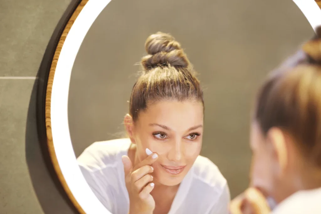 Woman looking in the mirror while applying facial cream to her cheek with a smile.