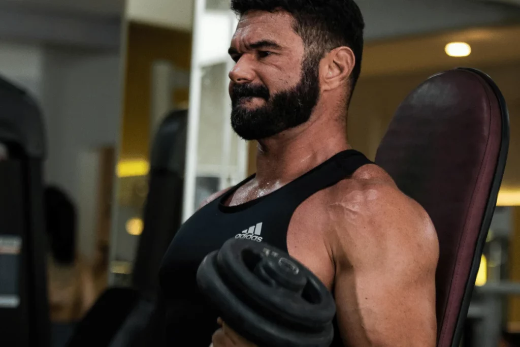 A muscular person with a black tank top lifts a dumbbell in a gym, showcasing their defined arms and serious workout focus.