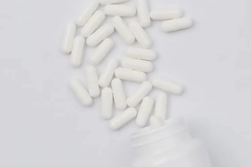 A scattered arrangement of white capsules beside an open bottle on a light gray background.