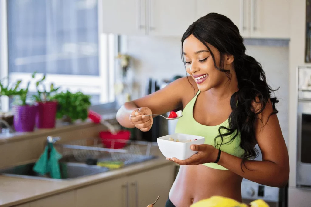 Smiling woman in a sports top enjoying a bowl of fruit in a bright kitchen, holding a spoon with a strawberry.