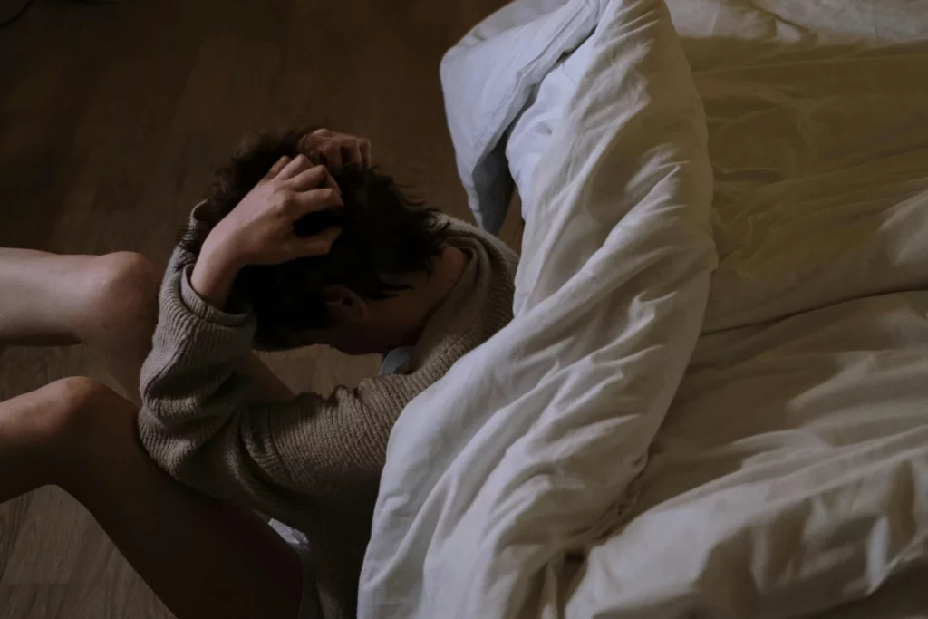 A person sits on the floor, clutching their head in distress, with a disheveled bed in the background, symbolizing emotional struggle.