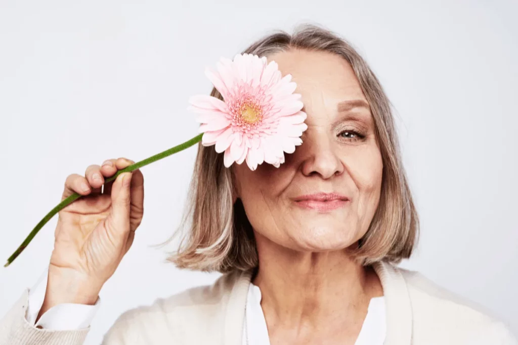 Mature woman with a pink daisy covering one eye, smiling softly.