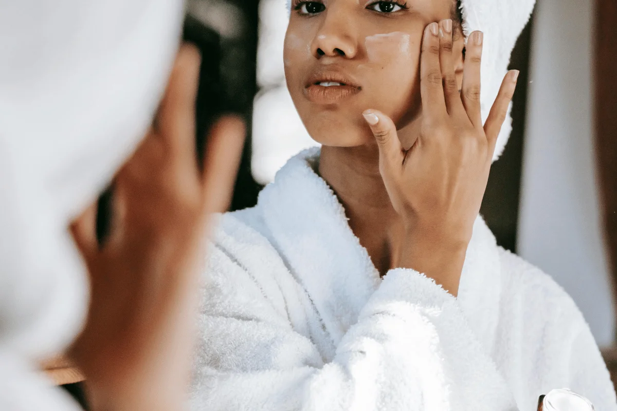 Woman applying face cream in front of a mirror, dressed in a white robe, for her skincare routine.