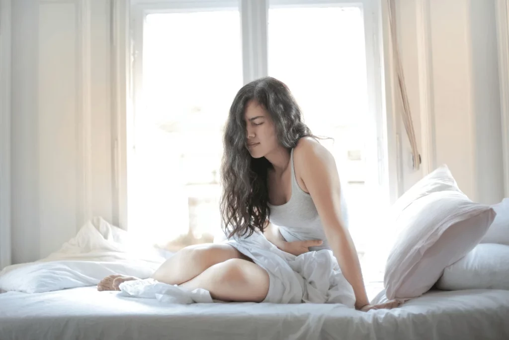 Woman sitting on a bed holding her stomach in discomfort.