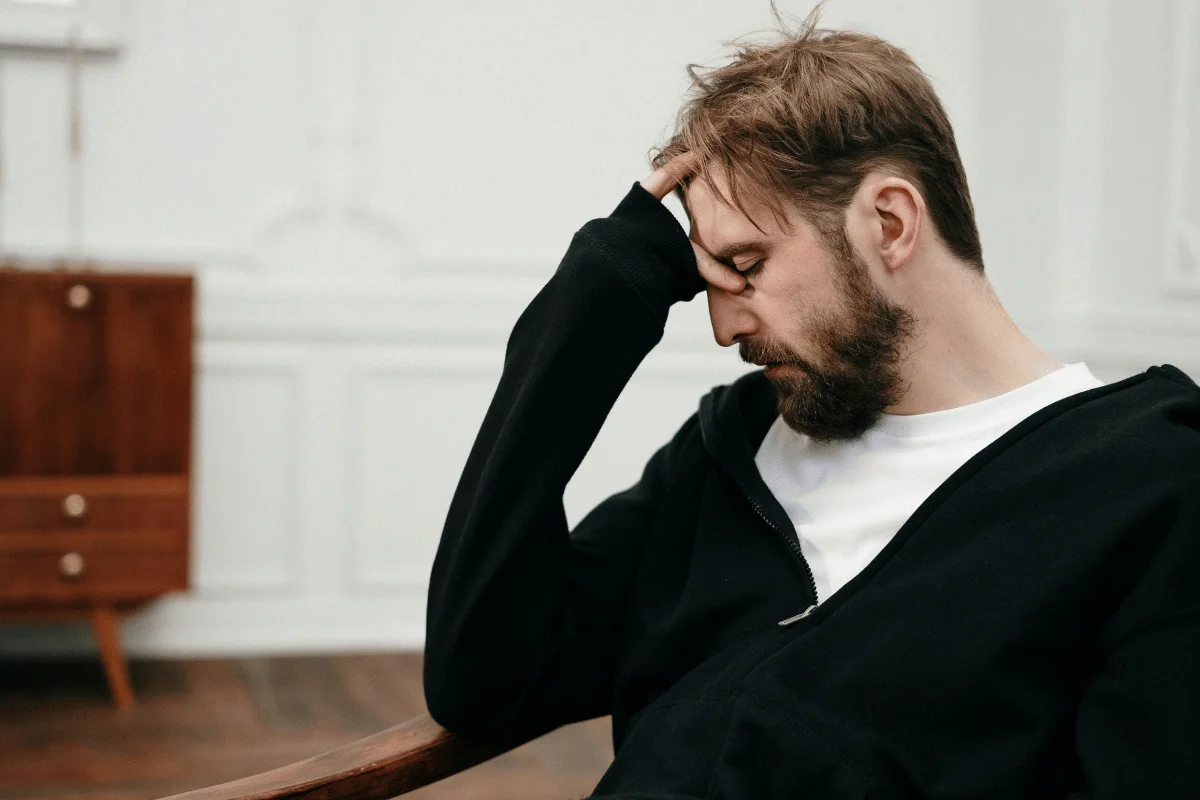 A person in a black hoodie sits with their head resting on their hand, appearing pensive, against a softly lit interior backdrop.