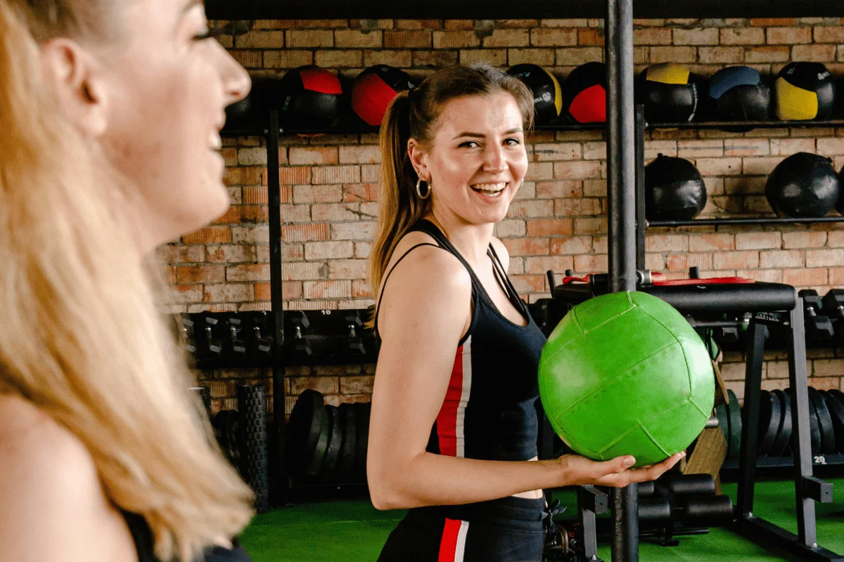 A woman in workout attire holds a bright green medicine ball in a fitness gym with weights and equipment in the background.