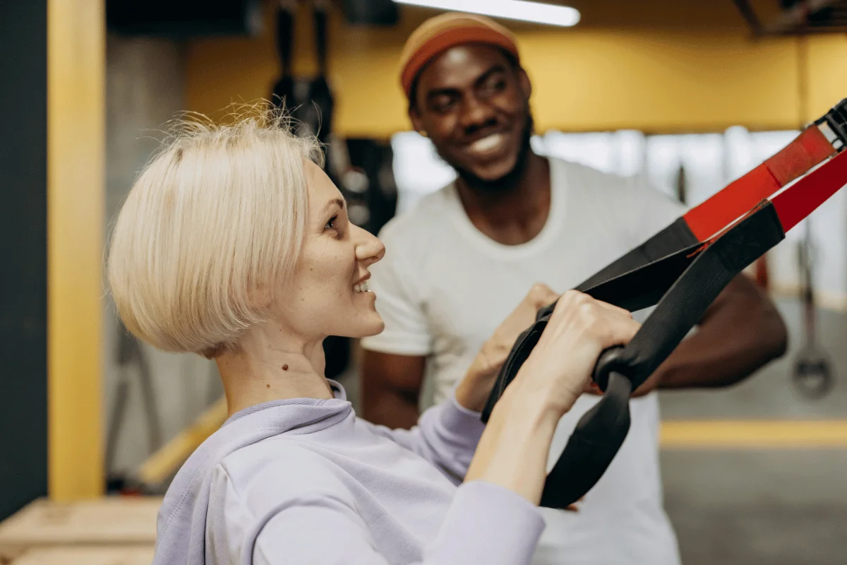 A woman with short blonde hair exercises using TRX suspension straps, supported by a trainer in a gym setting.