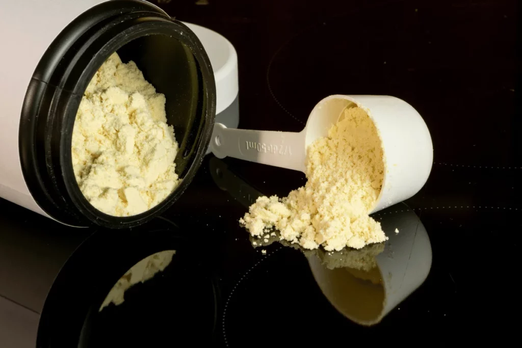 Close-up of a white container spilling yellow protein powder with a measuring scoop on a reflective surface.