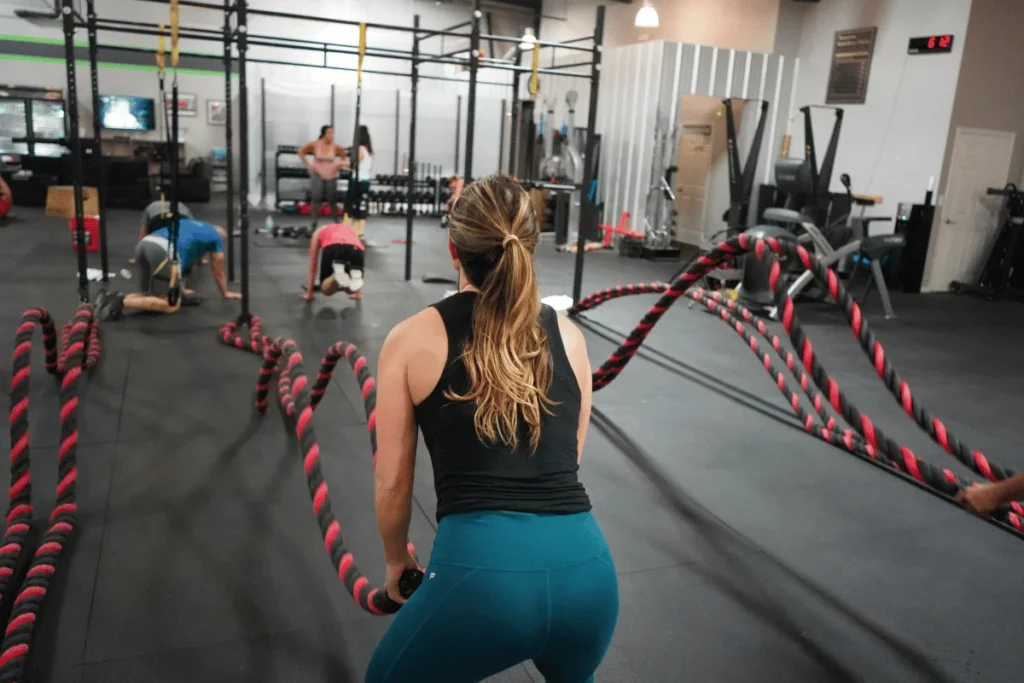 Woman performing battle rope exercises in a gym surrounded by other athletes in an intense training session.