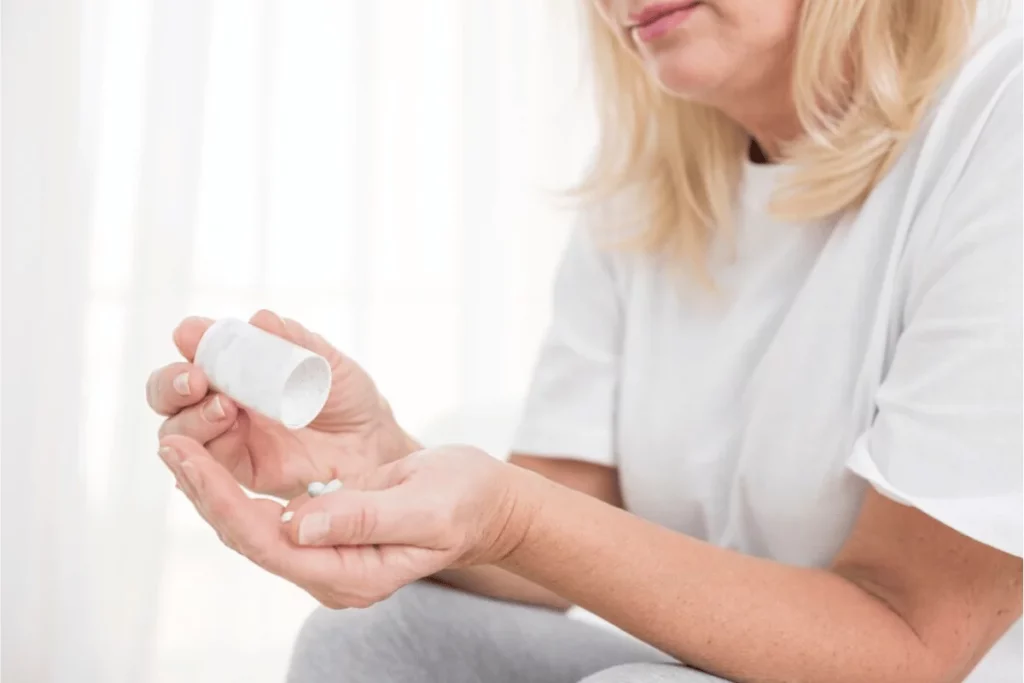 A middle-aged woman holds a white pill bottle with pills in her hand, contemplating medication, in a softly lit room.