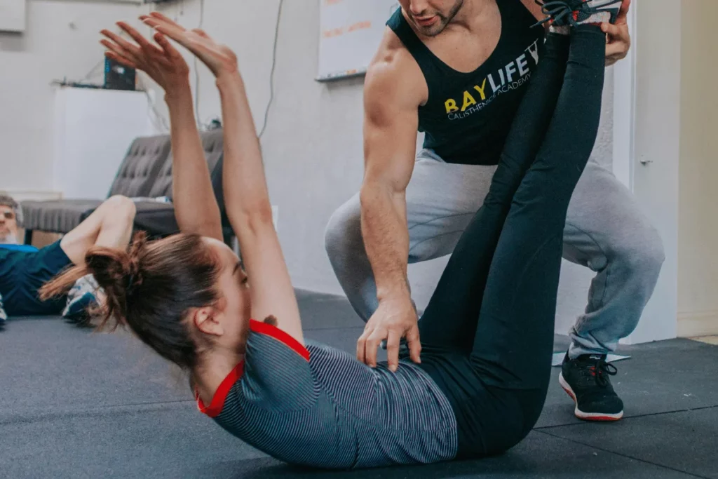 Fitness trainer assisting a woman with a stretching exercise in a gym.