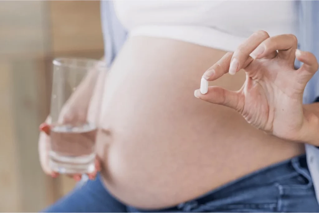 Pregnant woman holding a small white pill and a glass of water, preparing to take medication.