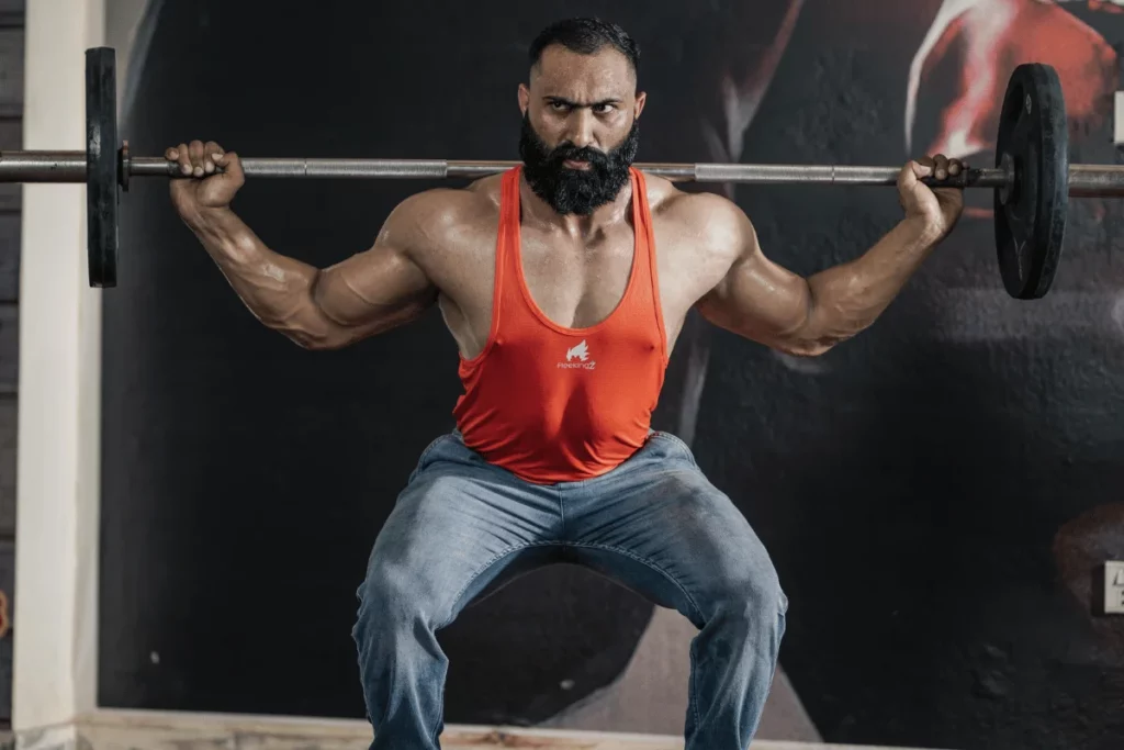 A muscular individual performs a squat with a barbell while wearing a red tank top and blue jeans in a gym setting.