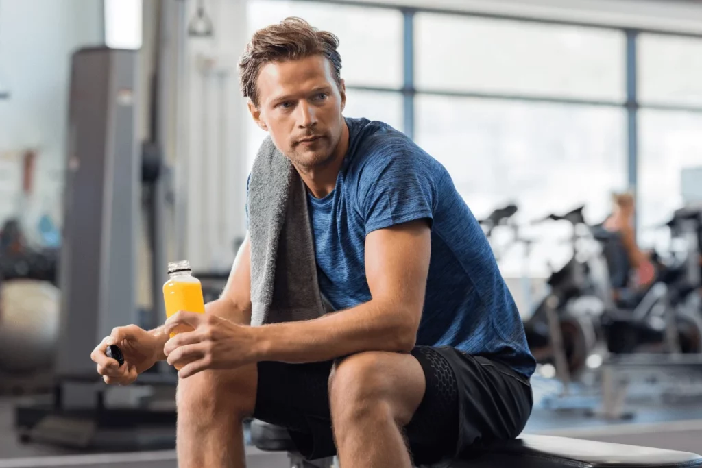 Man sitting in a gym holding a sports drink during a workout break.