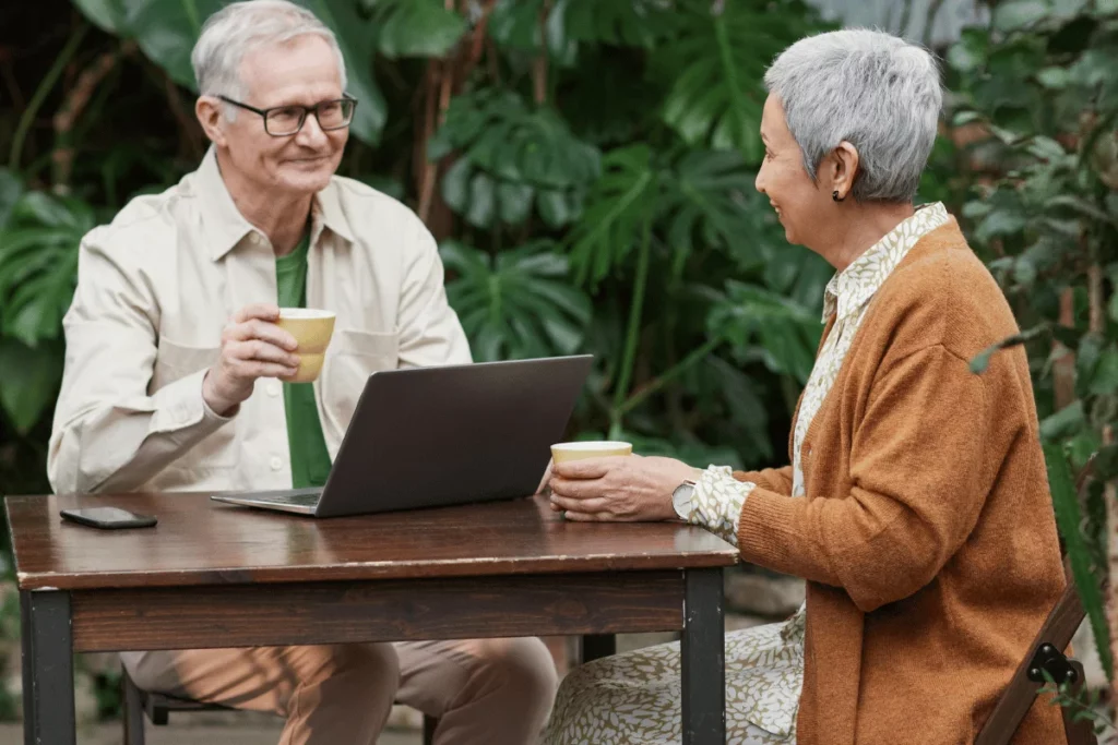 Two people sit at a wooden table in a lush garden, each holding a cup, while a laptop is open between them.