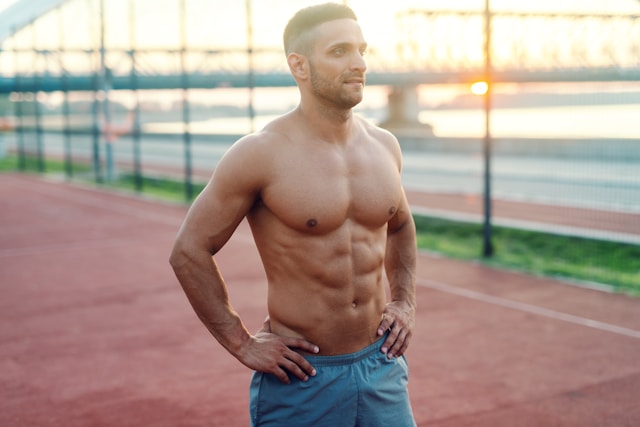 A shirtless athletic man with defined muscles poses on a running track during sunset.