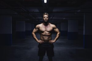 A shirtless muscular man poses with hands on hips in a dark gym environment.