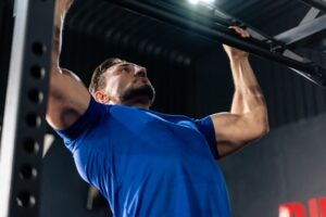 A bearded man in a blue shirt performs pull-ups on a gym exercise bar.