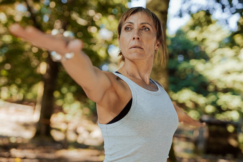 A middle-aged woman in a gray tank top extends her arm while exercising outdoors.