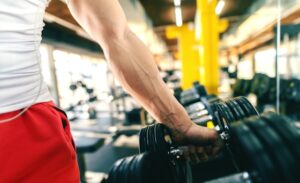 A person's muscular arm grips a black dumbbell from a weight rack in a bright gym.