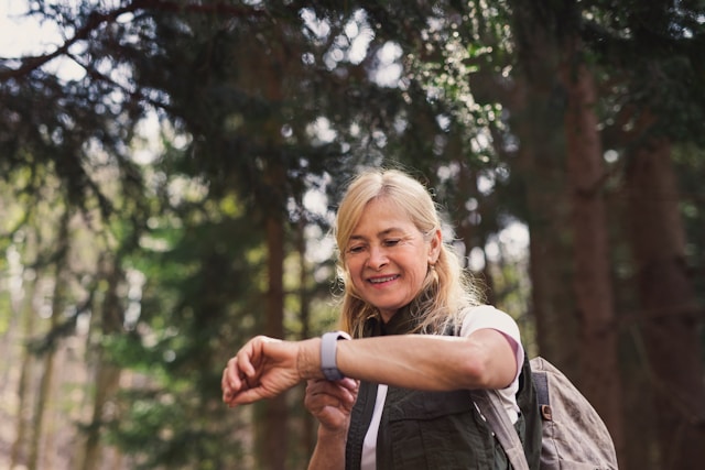A smiling blonde woman stretches her arms outdoors among trees while wearing athletic clothing.