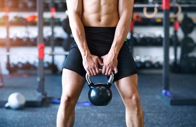 An athletic man holds a black kettlebell in a gym setting.