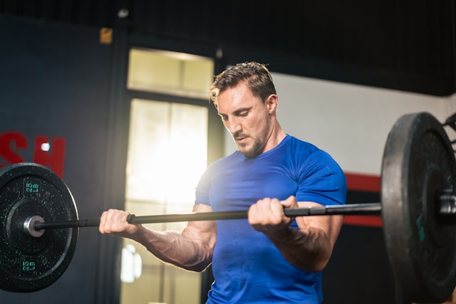 A man in a blue shirt performs a barbell curl exercise in a well-lit gym setting.