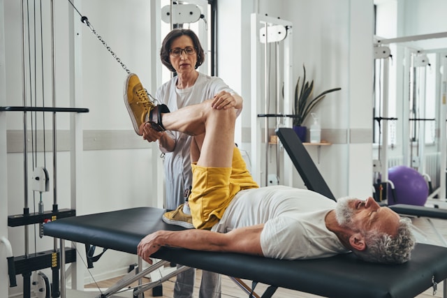 A female physical therapist assists an elderly man with leg exercises using pulley equipment in a rehabilitation facility.