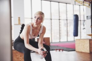 A blonde woman in workout clothes sits on a wooden box in a bright gym, holding her phone.