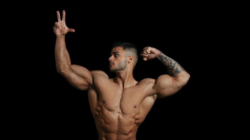 Muscular shirtless man with arm tattoo flexing biceps and striking a dynamic pose against a black studio background.