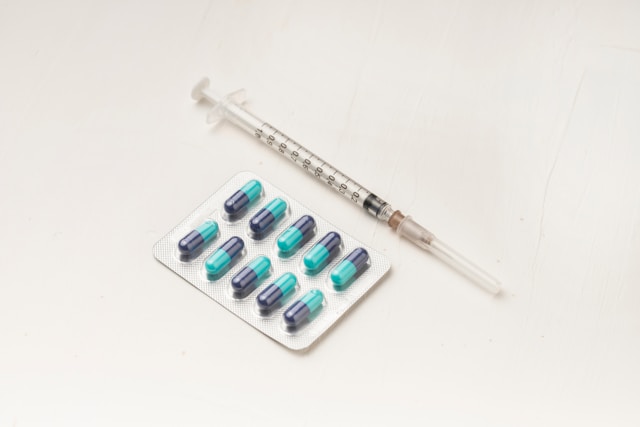 A medical syringe lies next to a blister pack of green and blue capsules on white background.