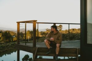 Bearded man in brown hoodie sits on wooden deck with cable railing, holding cup while overlooking scenic countryside at golden hour.