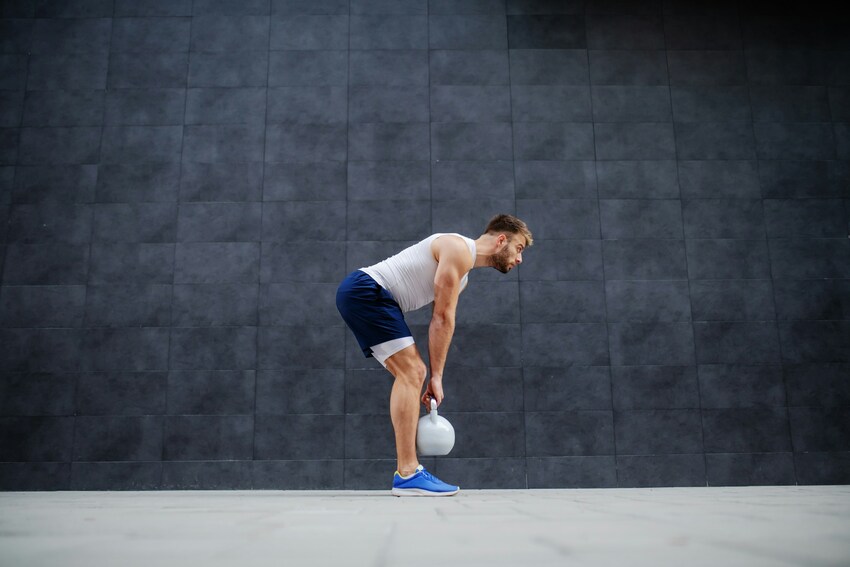 Athletic man in white tank top and blue shorts performing kettlebell deadlift exercise against dark tiled wall in gym.