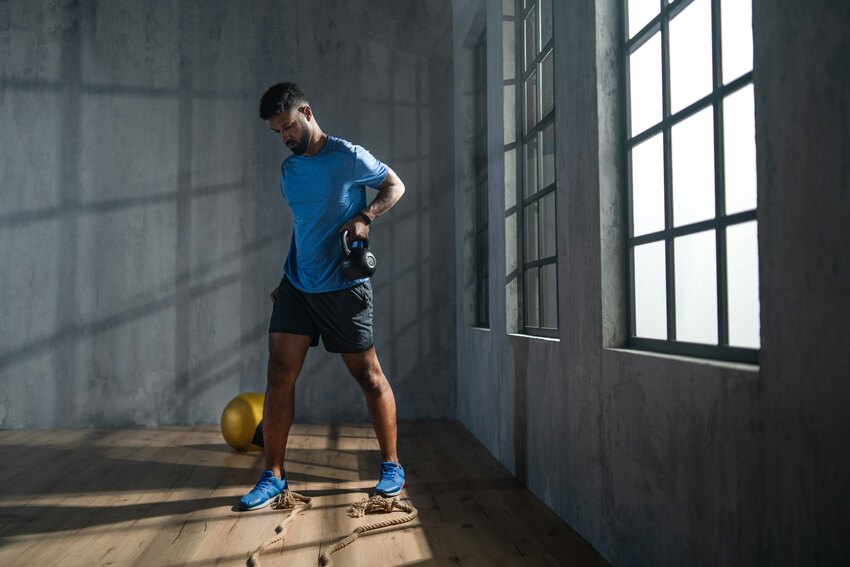 Man in blue athletic shirt exercising with dumbbell in industrial gym space with concrete walls, large windows, and wooden floor.