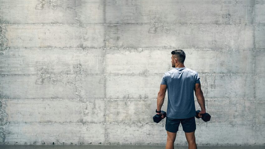 Man in gray athletic shirt holding dumbbells at his sides while facing weathered concrete wall in industrial gym setting.