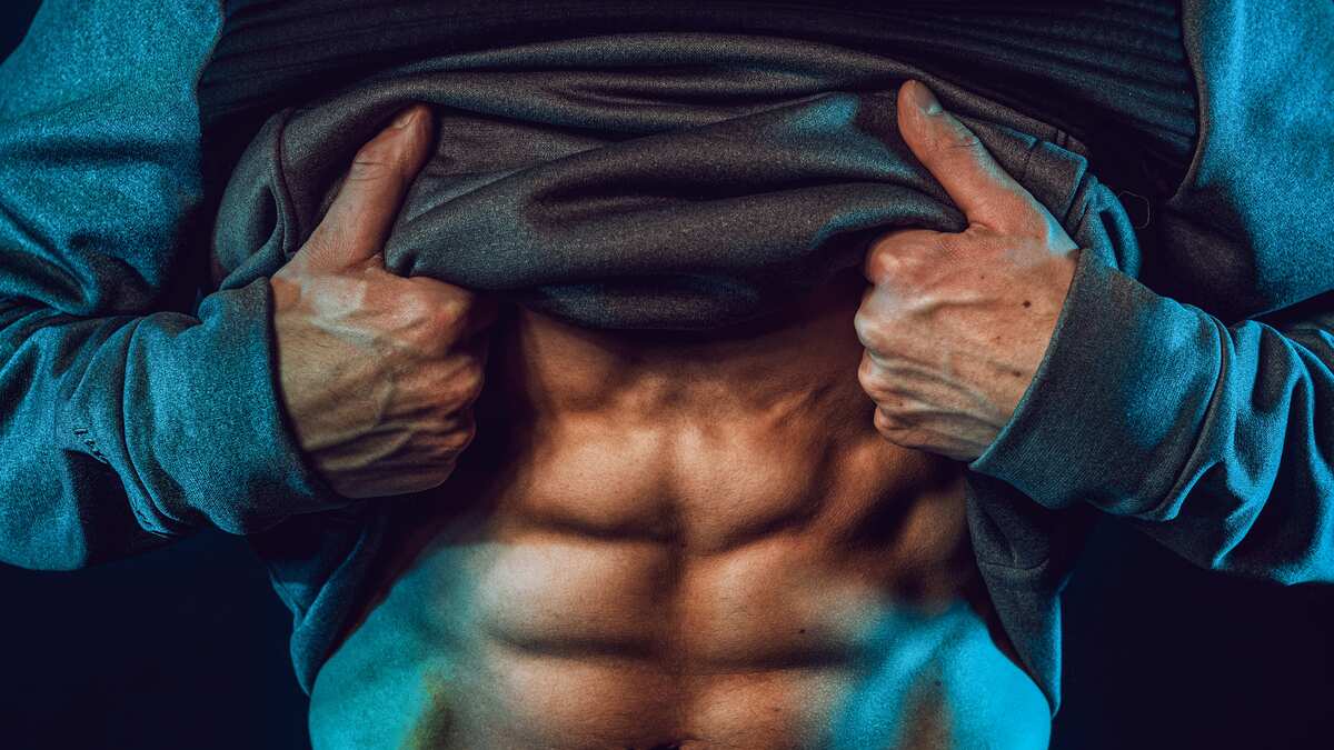 Muscular man in dark hoodie lifting shirt to reveal defined abdominal muscles in dramatic blue-tinted lighting for fitness photography.